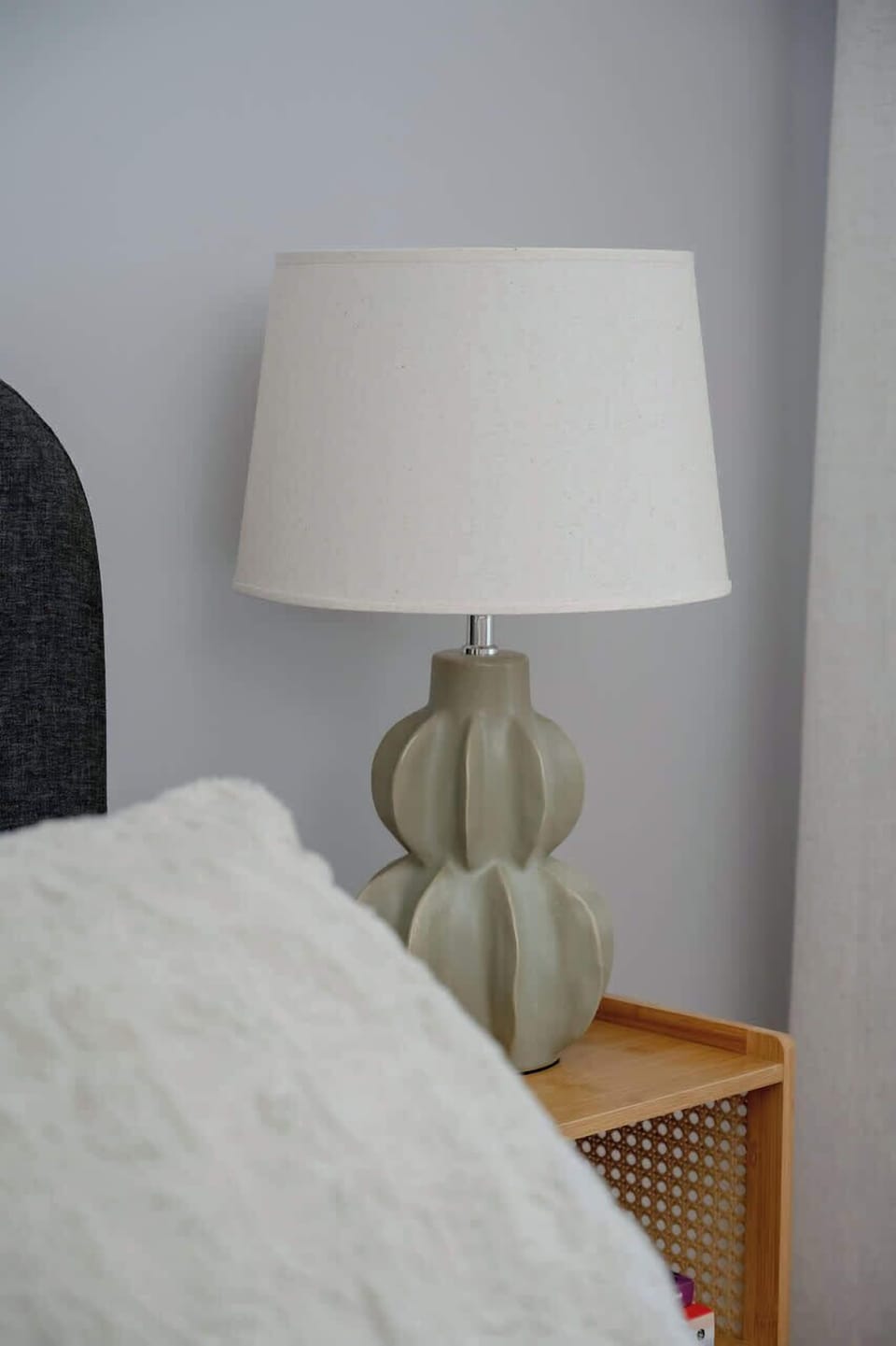 A bedside table with a textured ceramic lamp sits beside a neatly made bed. The setup is cozy and minimal, with soft lighting.