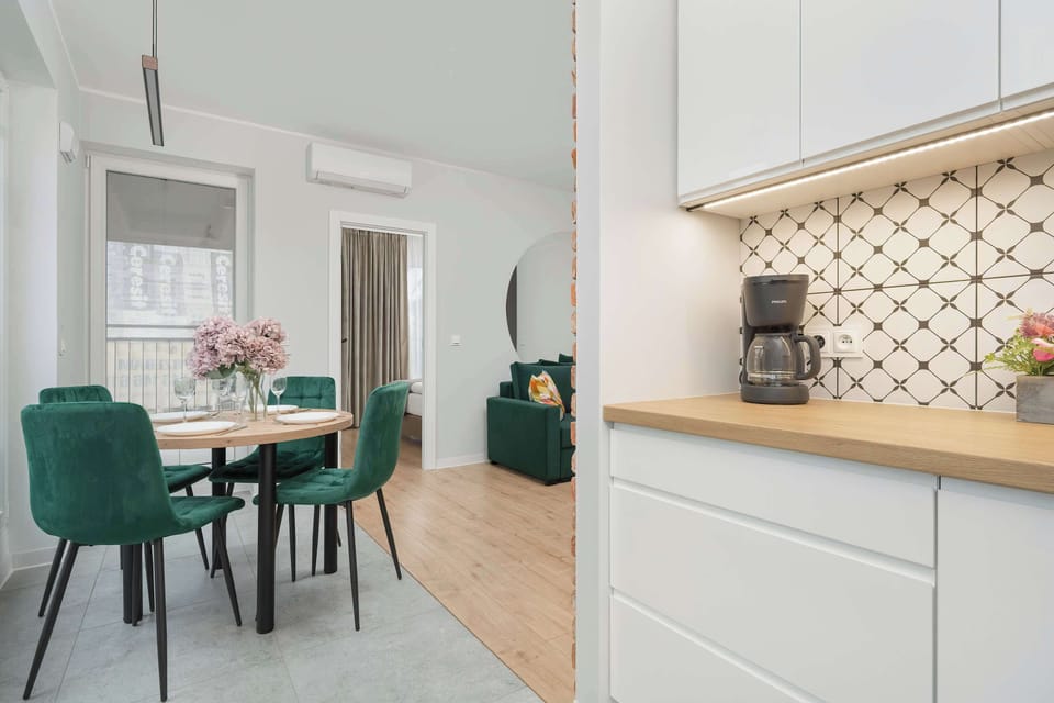 A dining table with a round top and green chairs creates a cohesive connection between the kitchenette and living room.