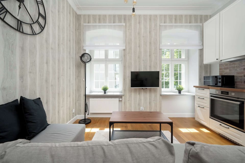 A modern living room corner with a sectional sofa, coffee table, flat-screen TV, and wall clock, all set against patterned wallpaper and large windows.