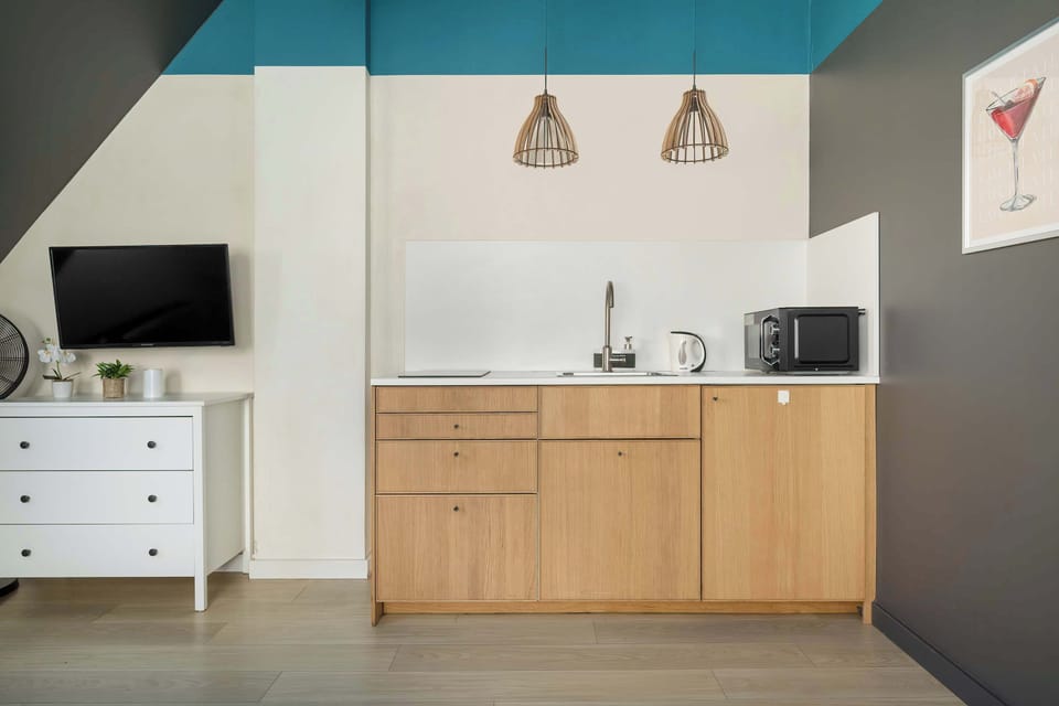 A view of a minimalist kitchen with wooden cabinetry and a white wall. The kitchen connects to a living area with a small dining table and stylish light fixtures on the ceiling.