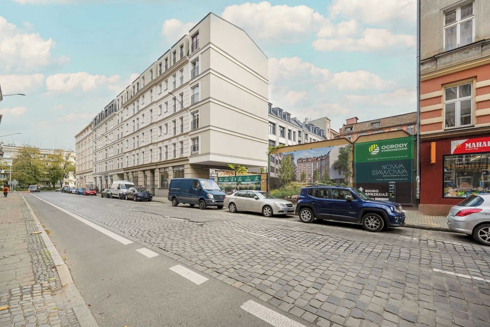 A street view showing a quiet urban scene, with both modern and older buildings. Several parked cars are visible along the road, and the atmosphere is calm and urban.