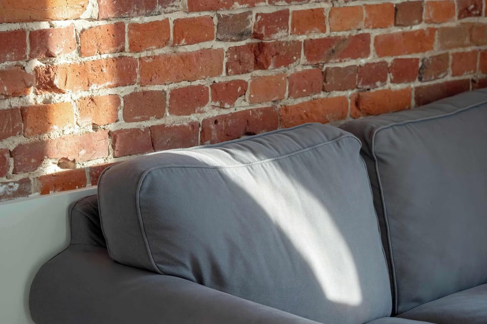 A close-up of the gray sofa against the brick wall, with natural light creating soft shadows. The exposed brick adds texture and warmth to the space.