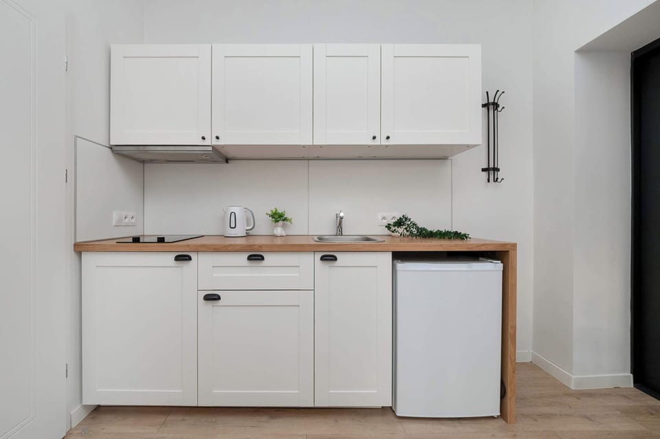A compact kitchen area featuring white cabinets and a small fridge. The setup is functional, with light wooden countertops and a small shelf for kitchen essentials.