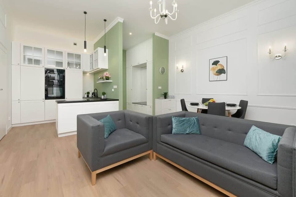Open-concept living room with a gray corner sofa, TV on a white-paneled wall, and a green accent leading to the kitchen.

