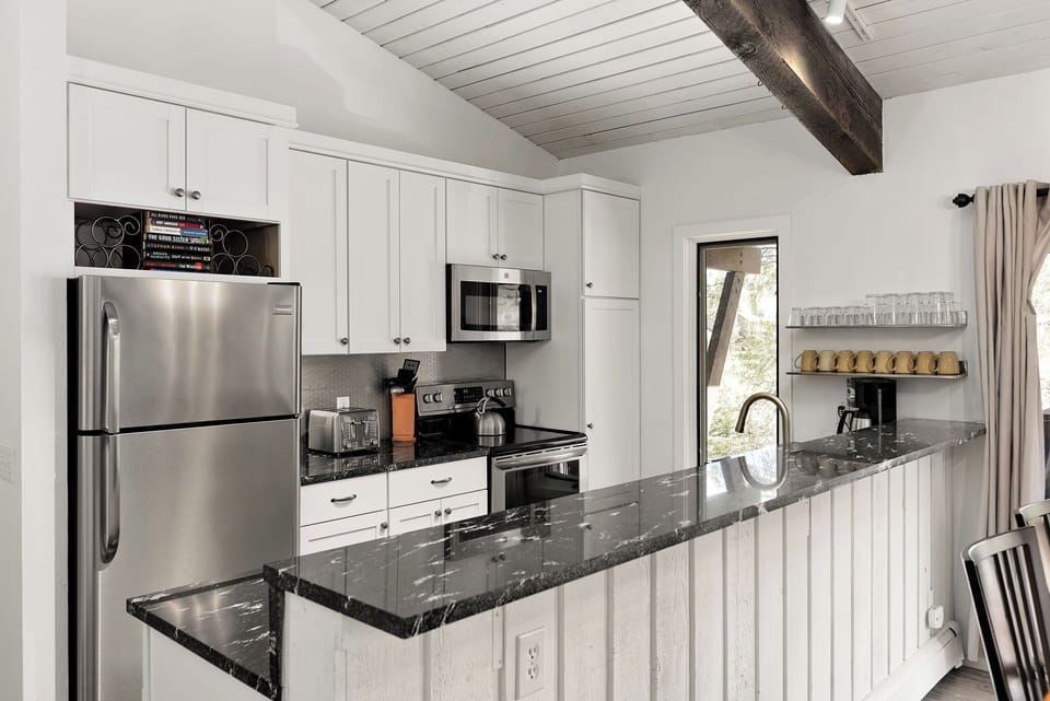 Modern kitchen with white cabinetry, stainless steel appliances, black granite countertops, and a white paneled island. The space features a vaulted ceiling with a wooden beam and a view of outdoors.