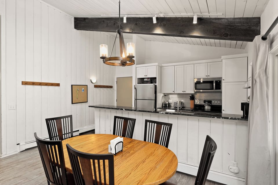 A dining area with a wooden table and six black chairs is next to a kitchen with white cabinets, stainless steel appliances, and a black countertop. The room features white paneled walls and a chandelier.