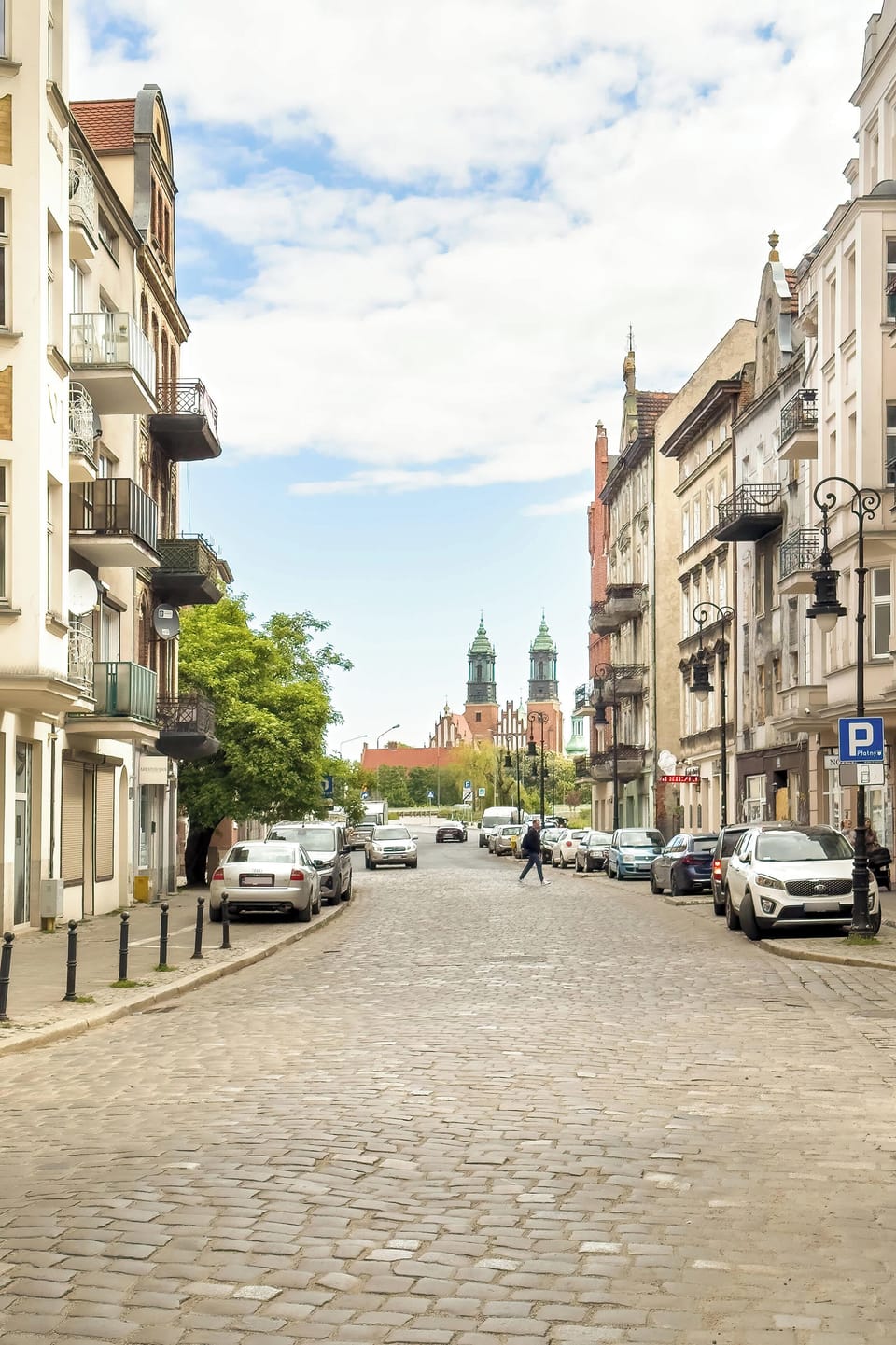 A picturesque cobbled street view with classic townhouses and a distant church tower under a bright sky, evoking local charm.

