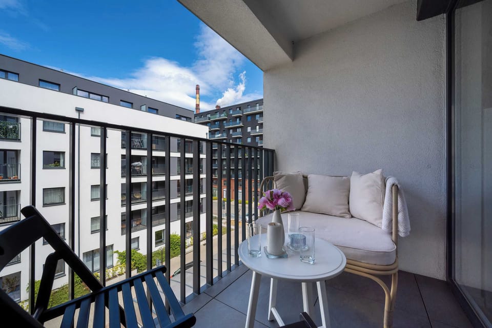 A private balcony is furnished with a comfortable white armchair and a small round table holding a bouquet of flowers. The view overlooks a neighboring modern residential building under a bright sky.