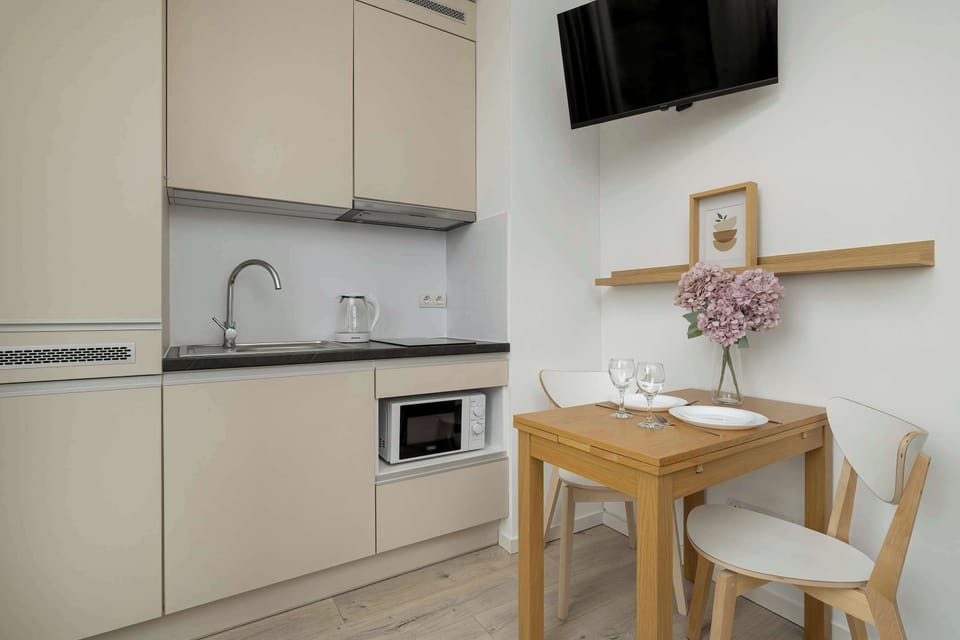 The kitchenette with light-coloured furniture matches the design of the entire flat interior. A flat-screen TV hangs above the wooden table.