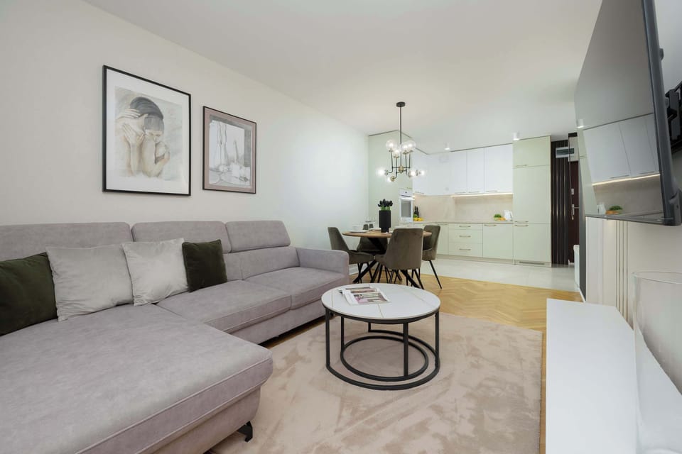 A bright living room with a gray sofa, a round coffee table, a TV on the wall, and large windows with curtains.