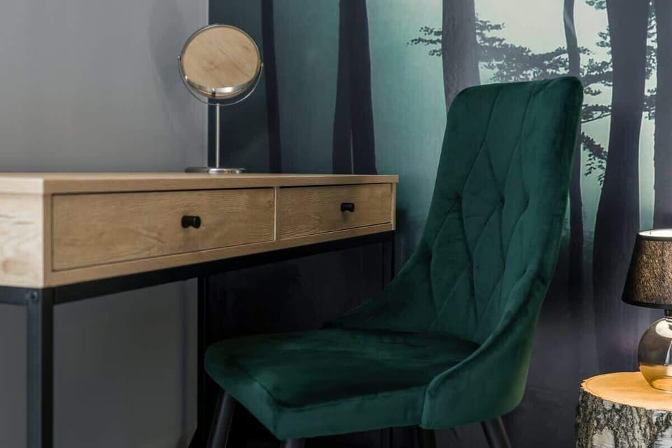 Workspace within a bedroom, including a wooden desk, green velvet chair, and a window with dark curtains for natural light.