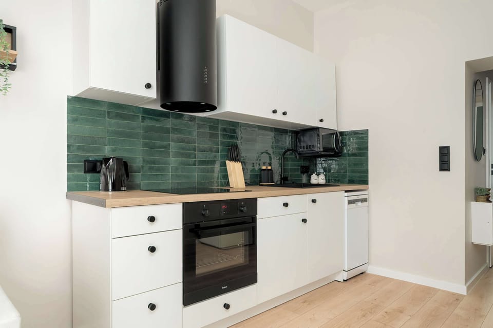 A modern kitchen with dark green tiles, white cabinets, and wooden countertops. Fully equipped with an oven, cooktop, and extractor hood.