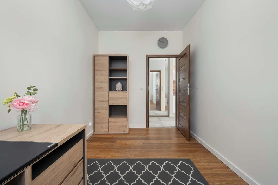 A minimalist interior with a wooden floor, a desk with flowers, perfect for remote work, a tall bookcase and an open door leading to another room.