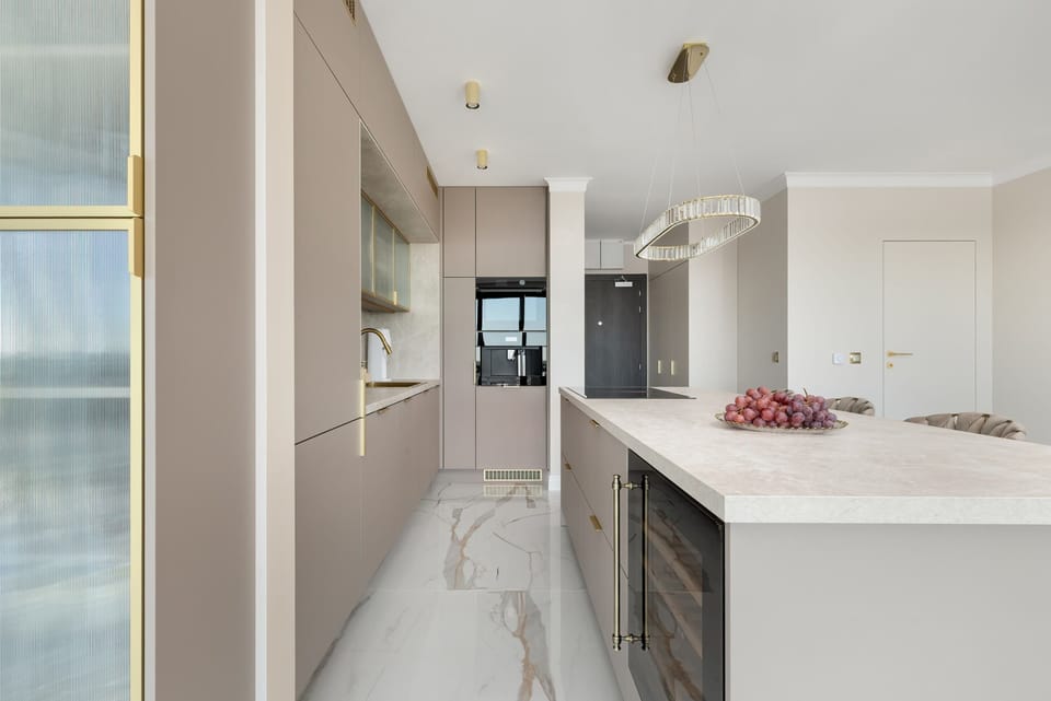 A modern kitchenette with an elegant island, marble floor and large floor-to-ceiling windows impresses with its spaciousness, natural light and stylish hockers with upholstered seats and golden legs, creating a luxurious space ideal for cooking.