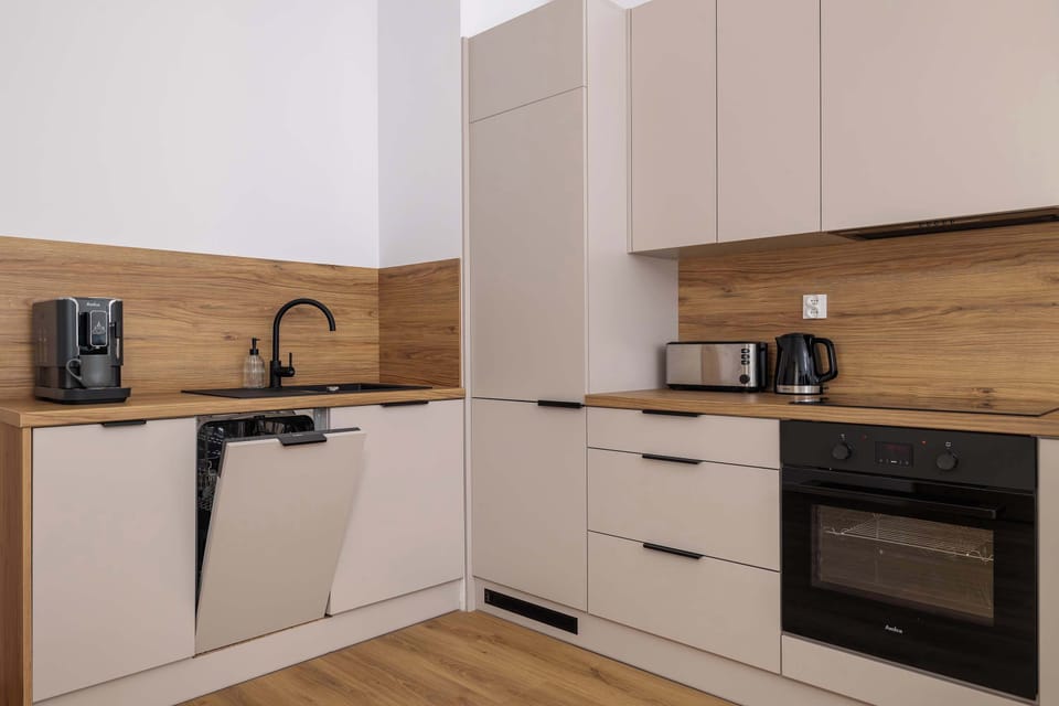 Modern kitchen with light beige cabinetry and wooden countertops, giving it a sleek and contemporary look. The kitchen features a built-in dishwasher with the door slightly open, a black sink with a matching faucet, and several small appliances.