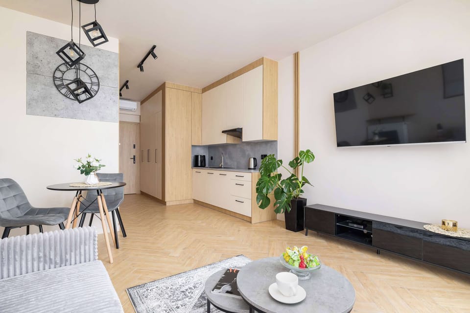 Studio with a gray sofa, Smart TV, complemented by warm wood flooring and tasteful decor. Kitchenette in the background.
