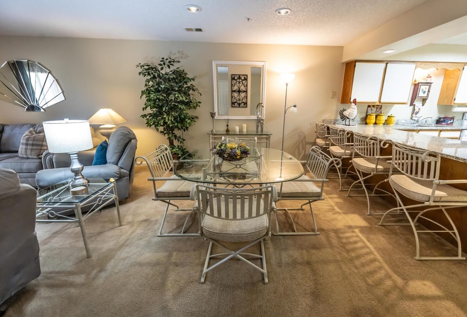Dining area with glass-top table, six cushioned metal chairs, beige carpeting, and adjacent granite breakfast bar with stools.