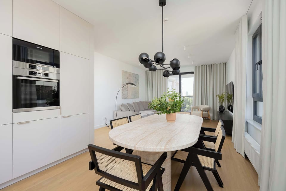 A bright dining area with a round table and black chairs. Open kitchen shelves and modern lighting complete the look.