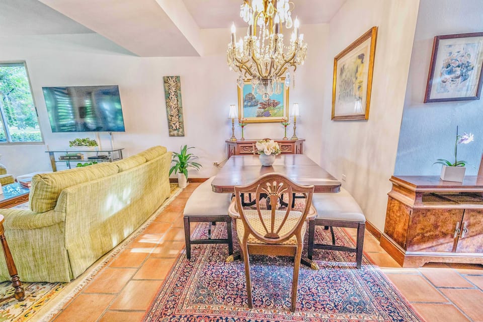 An open-concept living and dining area. The dining space features a wooden table with chairs and a bench under a prominent chandelier, while the living area has a large sofa and access to an outdoor space through sliding doors.