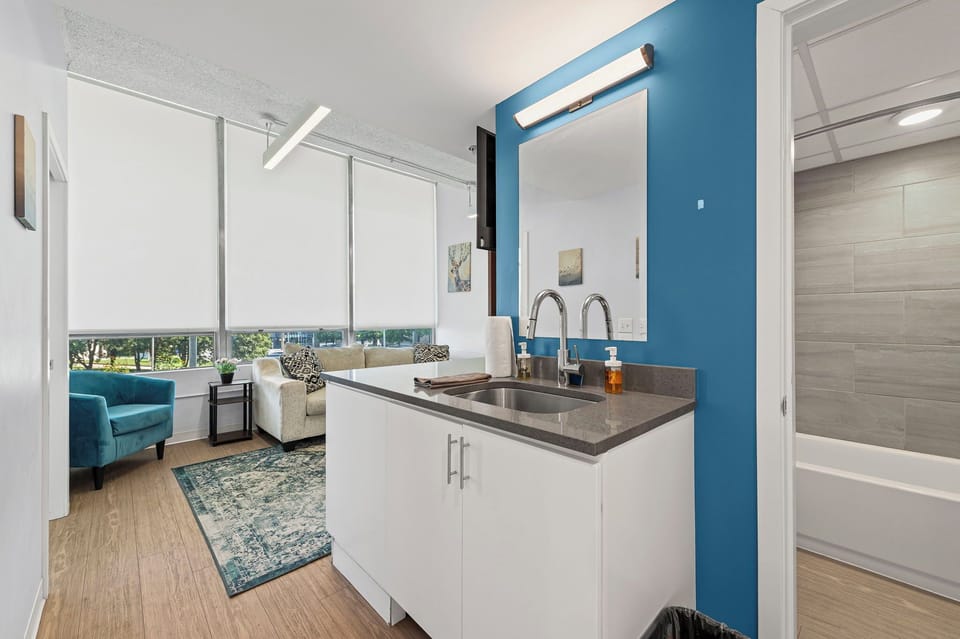 Vanity w/ bold blue wall, double faucets & mirror near bath & living area.