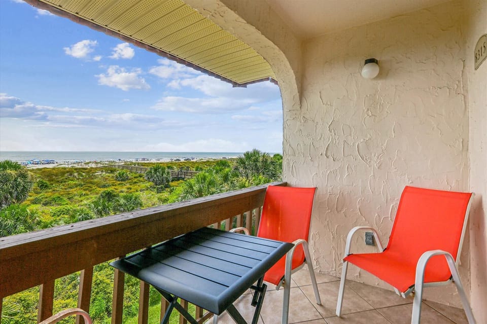 - Balcony with red chairs and table overlooks greenery and ocean horizon view  
- Peaceful outdoor setting perfect for morning coffee or evening drinks by the sea  
- Relax in coastal air with beautiful vacation scenery  
