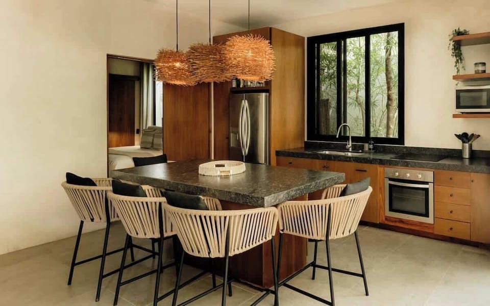 Dining area with wooden table, rope-wrapped chairs, dark countertops, and jungle view.
