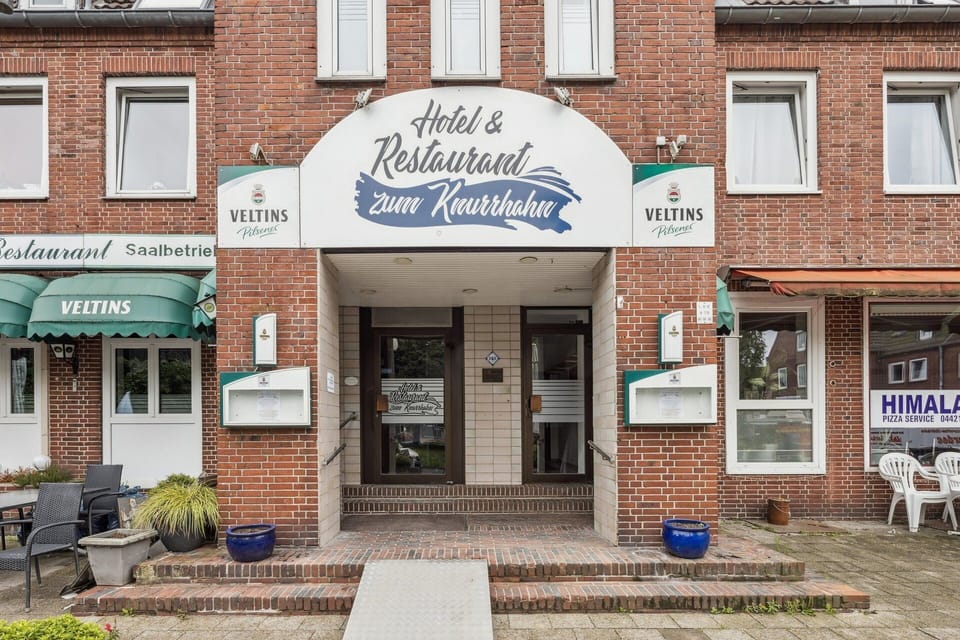Backsteinfassade mit überdachtem Hoteleingang und Restaurant „Zum Knurrhahn“ – zentraler, gut sichtbarer Zugang für Gäste.