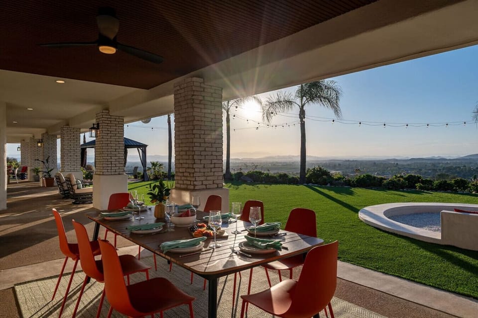 The covered patio offers ample seating, including a cozy conversation area around a propane fire pit coffee table, a round 4-person table with chairs, rocking chairs, an 8-person dining table, and a propane stainless steel grill.