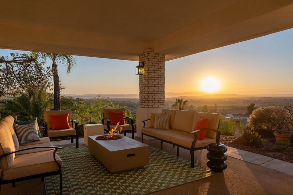 The covered patio offers ample seating, including a cozy conversation area around a propane fire pit coffee table, a round 4-person table with chairs, rocking chairs, an 8-person dining table, and a propane stainless steel grill.