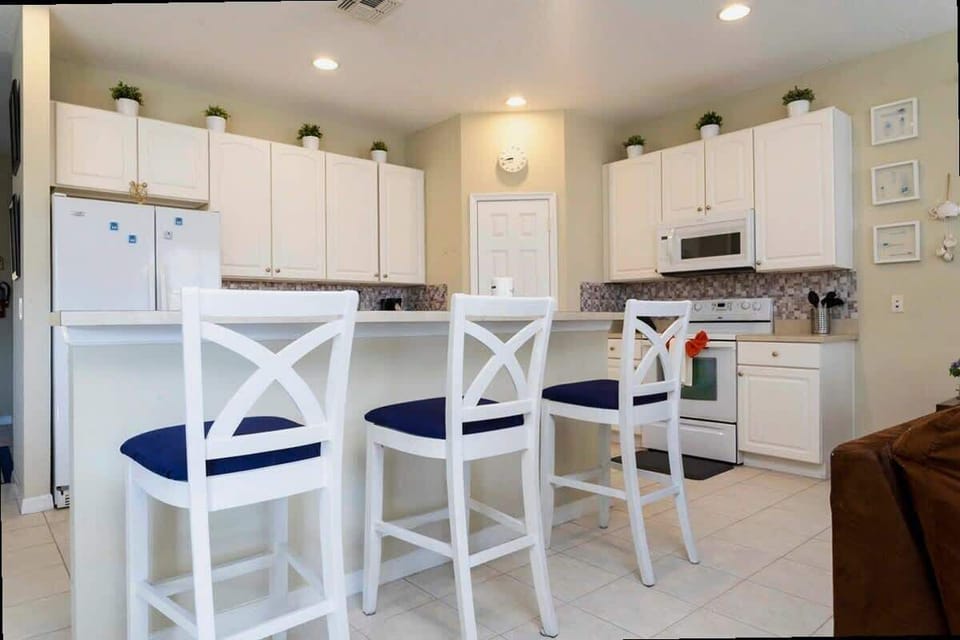 Bright kitchen with white cabinets, modern appliances, and a cozy breakfast bar with navy cushions on white chairs. Perfect for casual meals and socializing!