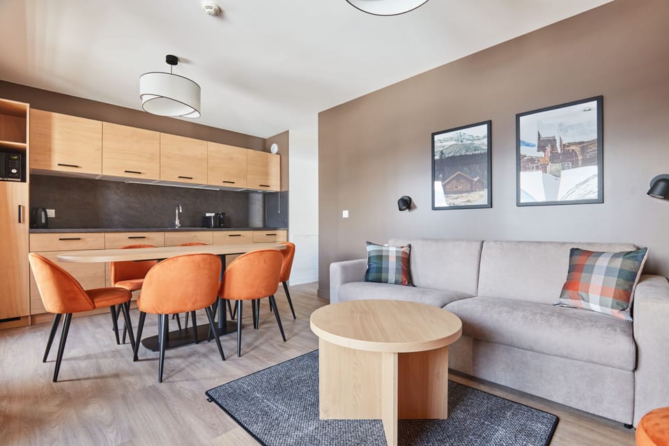 Living and dining area with round table, orange chairs, grey sofa, and a kitchenette with light wood cabinetry and dark countertop.