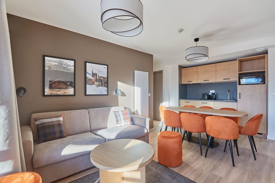 Living and dining area from another angle, with natural light through the large window and orange chairs around the table.