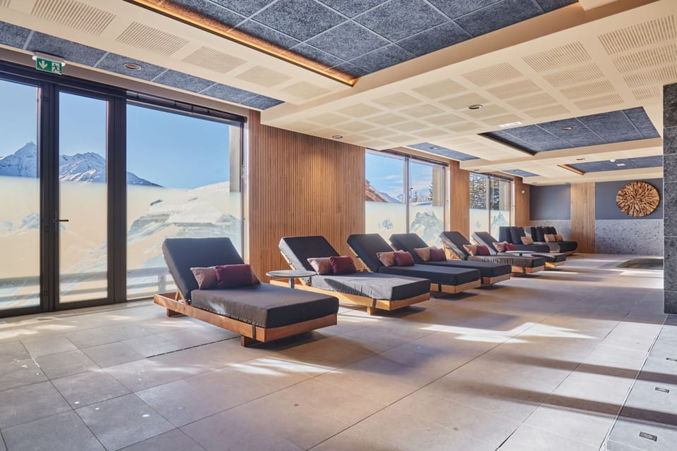 Grey lounge chairs with purple cushions line the poolside, with snow-covered peaks visible through the panoramic windows.