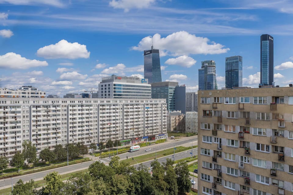 A cityscape view from the balcony, showing residential buildings and a skyline with high-rise towers under a bright blue sky.