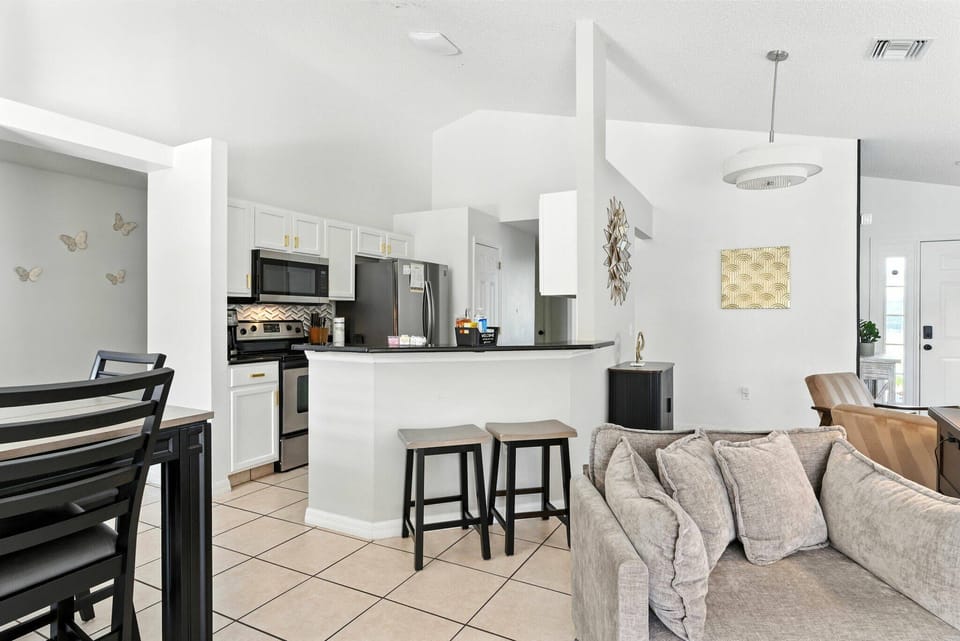 This bright, open-concept kitchen and living area boasts sleek stainless steel appliances and a cozy sofa for relaxation, with light tile flooring that enhances the spacious feel and stylish bar stools for casual dining.