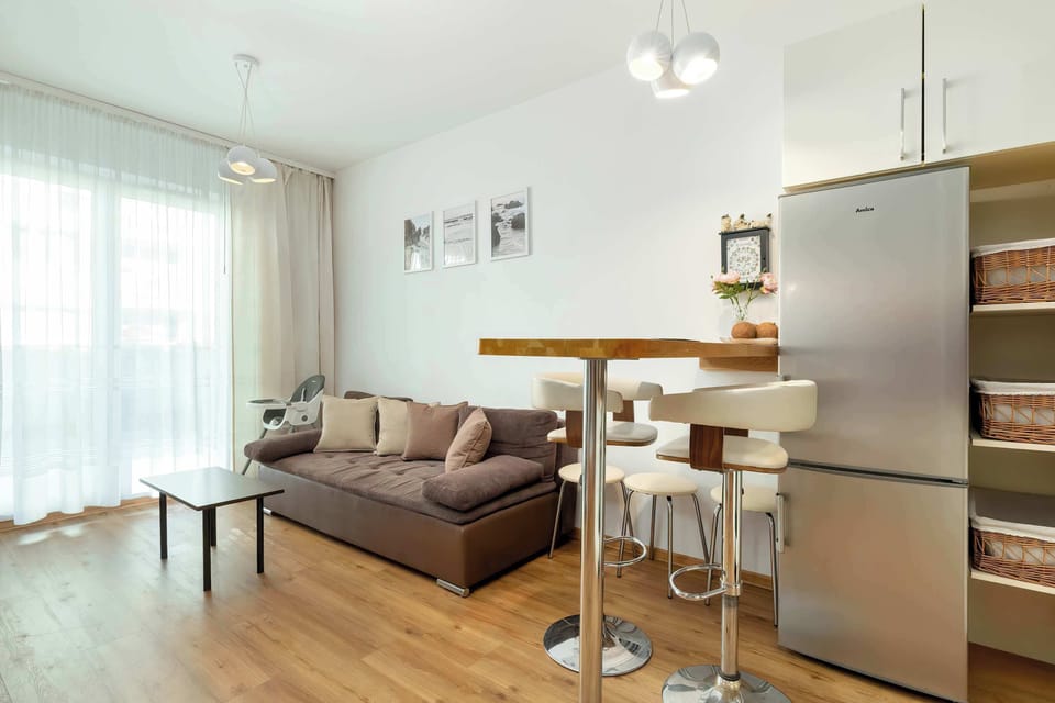 A modern studio interior featuring a sofa, mirrored wardrobe, and a kitchenette in a bright open space.