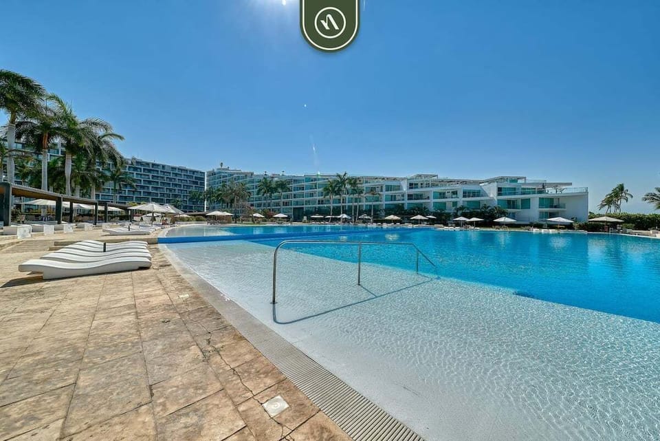 A stunning view of the expansive resort pool under a clear sky. The shallow entry point with a sleek handrail makes it easy to wade into the refreshing water, while the modern resort architecture provides a luxurious backdrop.