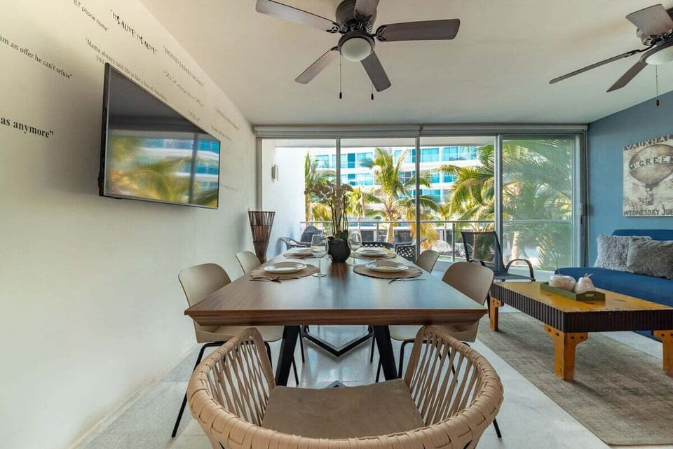 An expansive view from the dining table looking toward the terrace, highlighting the ceiling fans, wall-mounted television, and the natural light flowing in from the balcony.