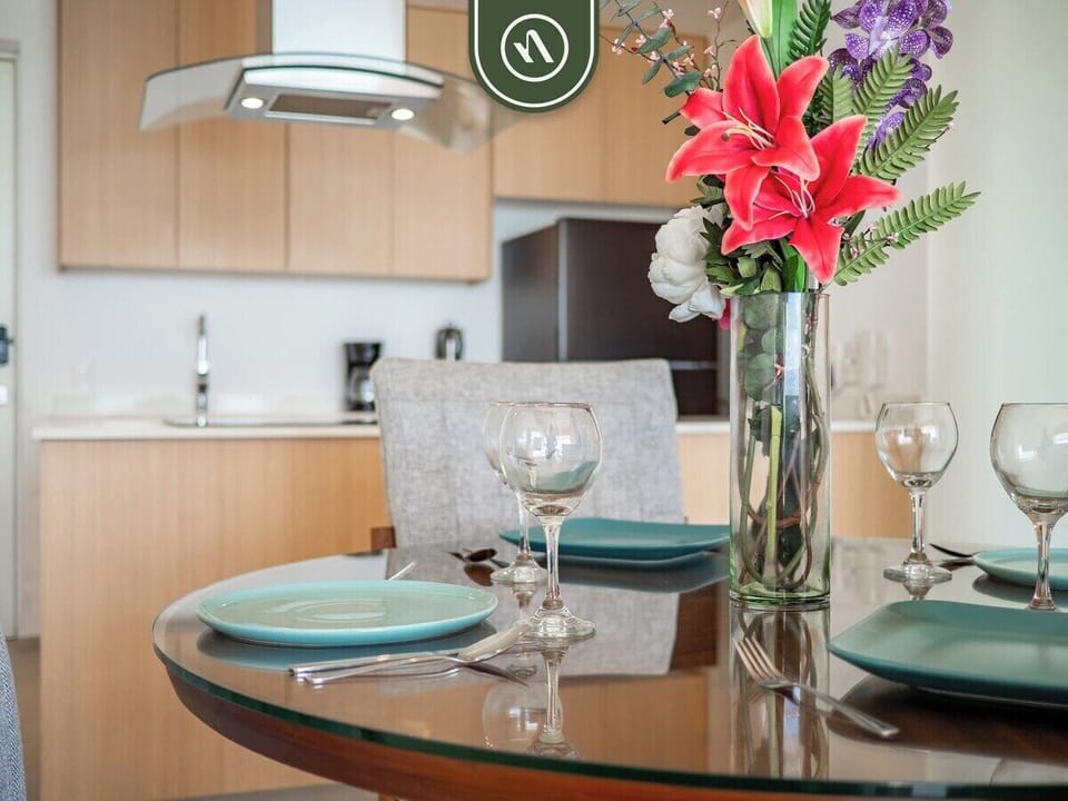 Enjoy a meal at the glass-top dining table. The adjacent kitchenette is fully equipped with modern appliances, light wood cabinetry, and stone countertops.