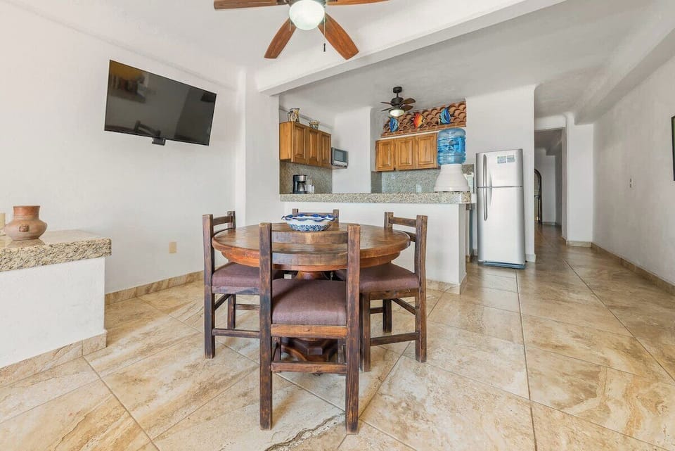 Open-concept room showcasing a round wooden dining table set beneath a wall-mounted TV, looking toward the kitchen counter.