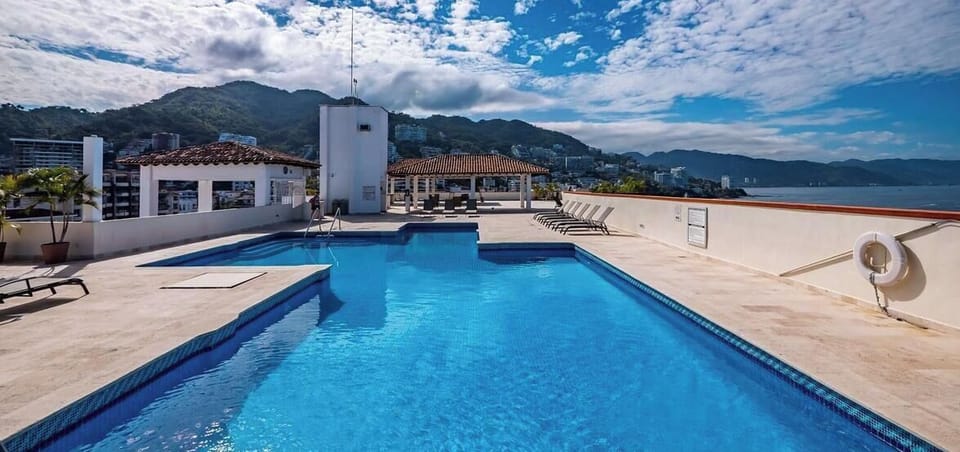 A wide view of the sparkling rooftop swimming pool surrounded by a spacious sun deck, under a dramatic cloudy sky.