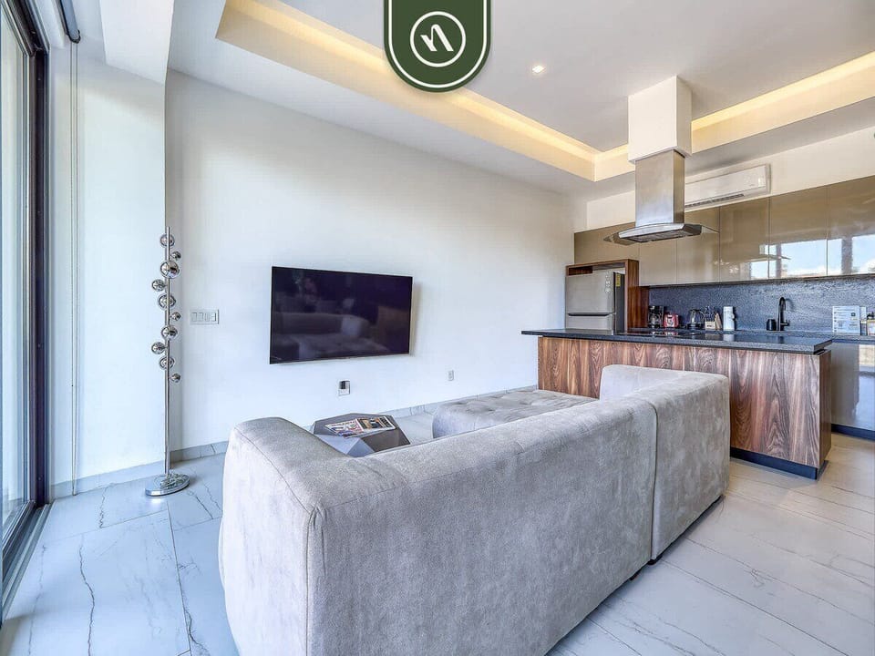 Bright living room featuring a wall-mounted TV, comfortable sofa, and kitchen view.