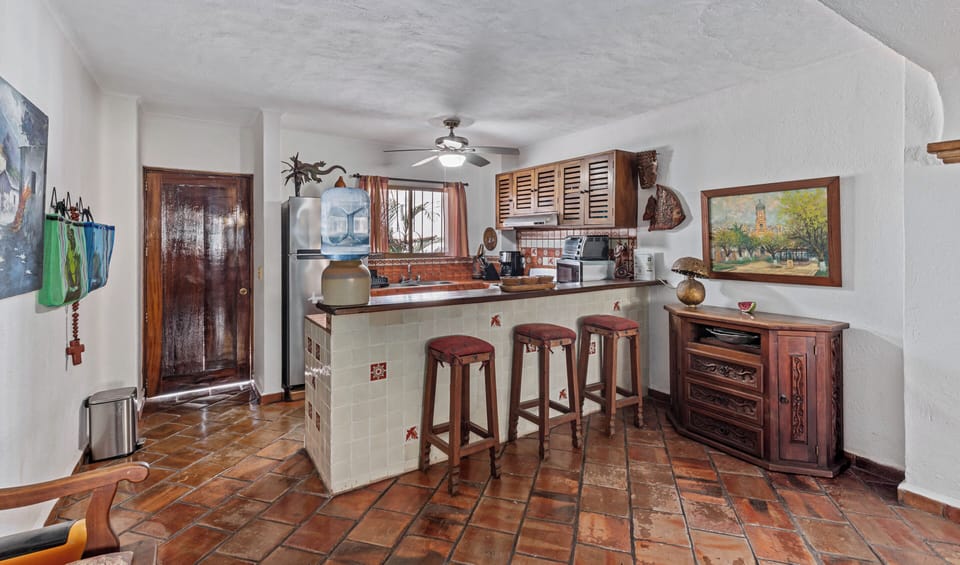 A rustic kitchen space featuring terracotta tile floors, traditional louvered wooden cabinetry, and a tiled breakfast counter with wooden barstools.