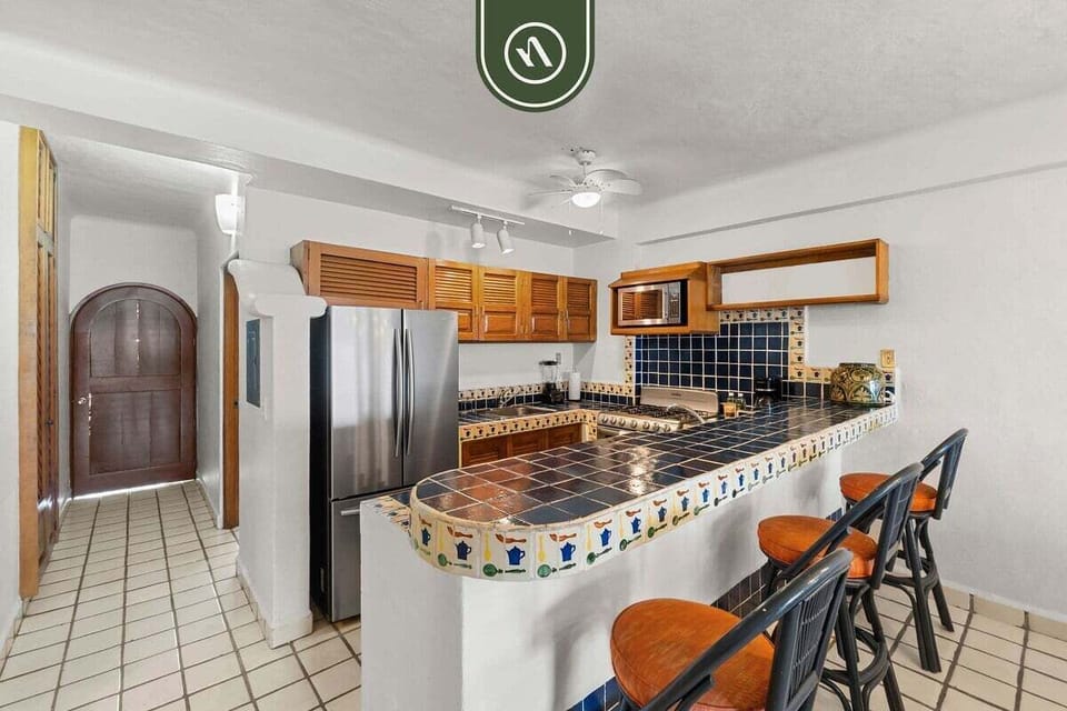 Classic Mexican-style kitchen featuring tiled countertops, wooden cabinetry, stainless steel appliances, and a curved breakfast bar with stools.