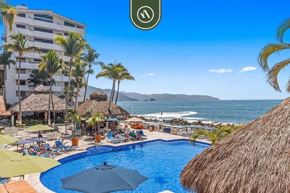 Sparkling blue swimming pool area bordered by palm trees and palapa structures right beside the ocean.