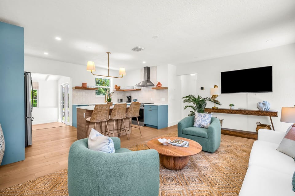 Elegant, sun-filled living room featuring designer coastal décor, plush swivel chairs, a chic white sofa, and custom wood accents, with tropical views and a seamless flow into the gourmet kitchen—perfect for luxurious relaxation and entertaining.