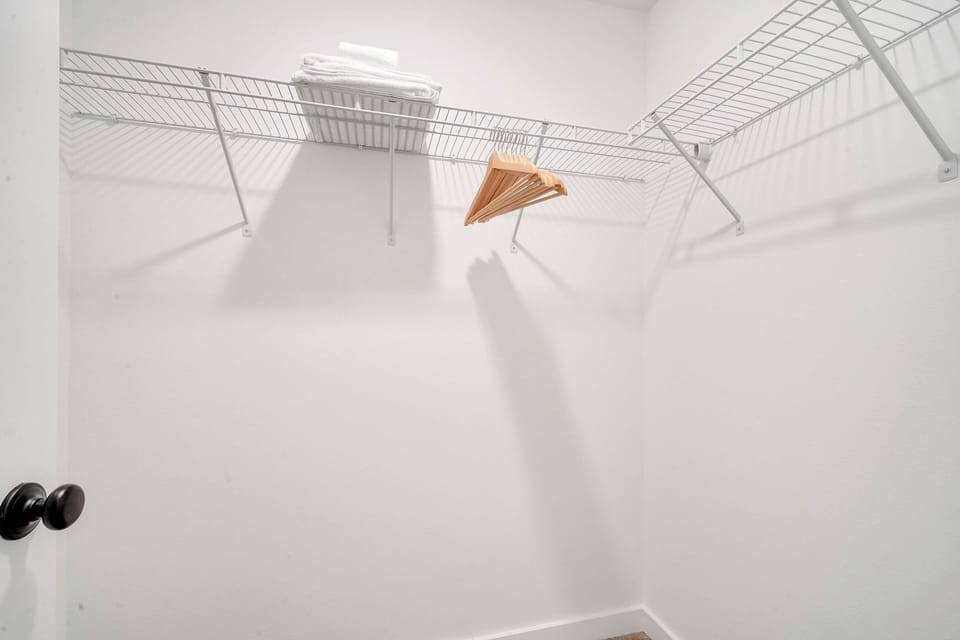 This spacious closet features sleek wire shelving and a wooden hanger, providing an organized space for your belongings. The bright, white walls add to the room's airy feel, creating a clean and simple atmosphere for your comfort.