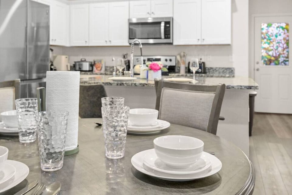 Stylish dining table set for four with glassware, dinnerware, and a paper towel holder. In the background, a modern kitchen features granite counters, stainless steel appliances, and a bright floral centerpiece, creating a warm and inviting space.