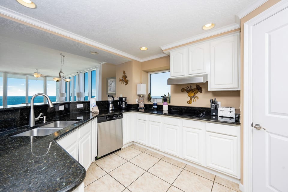 This kitchen features sleek black granite countertops, white cabinetry, and a stunning beachfront view through large windows