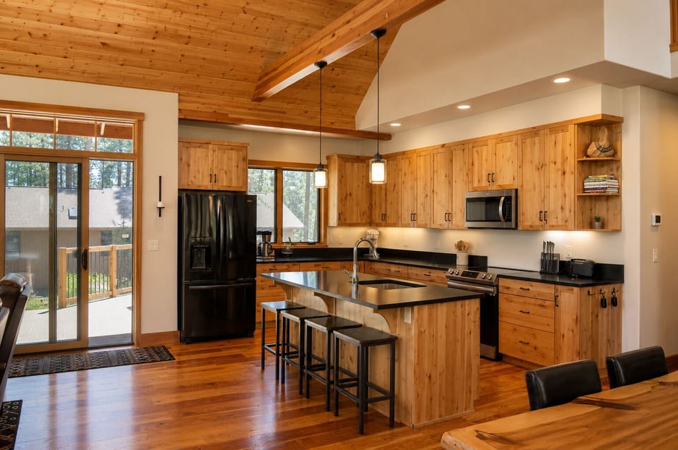 Your gourmet kitchen awaits with a spacious island, stainless steel appliances, and warm wood finishes throughout. Sunlight streams through large windows as you prepare meals in this inviting space with vaulted ceilings and modern amenities.